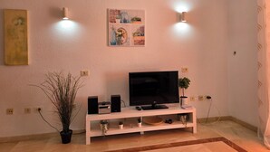 A living room with a flat-screen TV on a white entertainment unit, two wall-mounted lights, a painting, and a potted plant. - Casa Teide, ground floor Parque Santiago 1, heated pool, 100 m to sea+beach (Playa de la Américas)