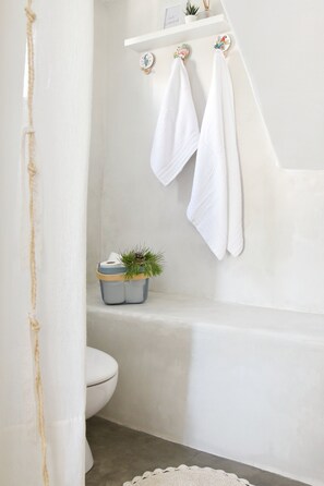 A bathroom with a bathtub, a toilet, and a shelf with towels and a small plant. - Koukounari Cottage Paros, Apartment With sea View (Paros)