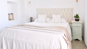A neatly made bed with a striped blanket, a bedside table with a plant, and a wall-mounted shelf with books. - Koukounari Cottage Paros, Apartment With sea View (Paros)
