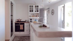 A modern kitchen with white cabinetry, a central island, and a window with curtains. - Koukounari Cottage Paros, Apartment With sea View (Paros)