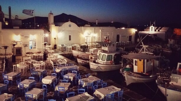 A marina with boats docked and tables set up for dining, a white building with a cross on top, and a flag flying. - Koukounari Cottage Paros, Apartment With sea View (Paros)