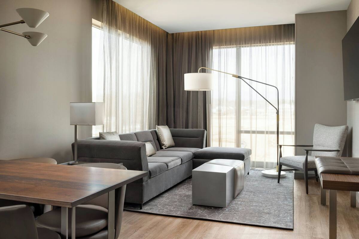 A modern hotel room with a sectional sofa, a small table, a lamp, and a view of the city through large windows.