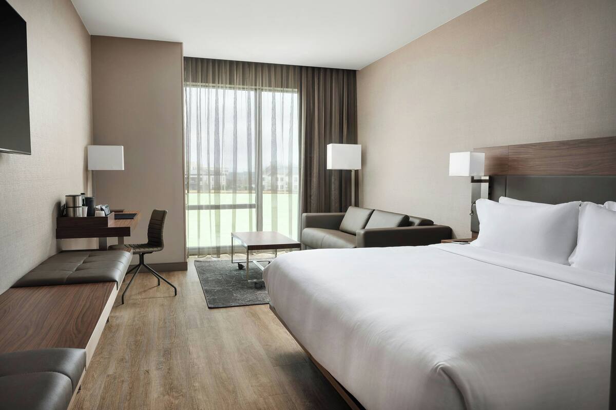 A modern hotel room with a large bed, a sofa, a desk, and a chair. There are two lamps on either side of the bed.