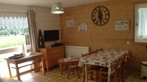 Una stanza accogliente con una parete in legno, una grande orologio, un tavolo da pranzo con sedie, una televisione e una finestra con tende.