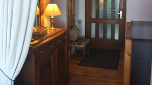 Un pasillo con suelo de madera, una puerta de madera con paneles de vidrio, una mesita con lámpara y una cortina blanca.