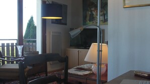 Una sala de estudio con un escritorio de madera, una computadora portátil, un cuaderno y un teléfono inteligente. Hay una silla, una lámpara y una ventana con vista a árboles.