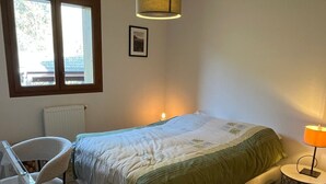 Ein Schlafzimmer mit einem Bett, einem Sessel, einem Nachttisch mit Lampe, einem Heizkörper und einem Fenster mit Blick auf ein Gebäude.