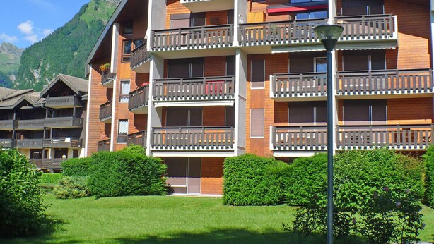 Un hotel a più piani con balconi, uno sfondo montuoso e un prato curato.