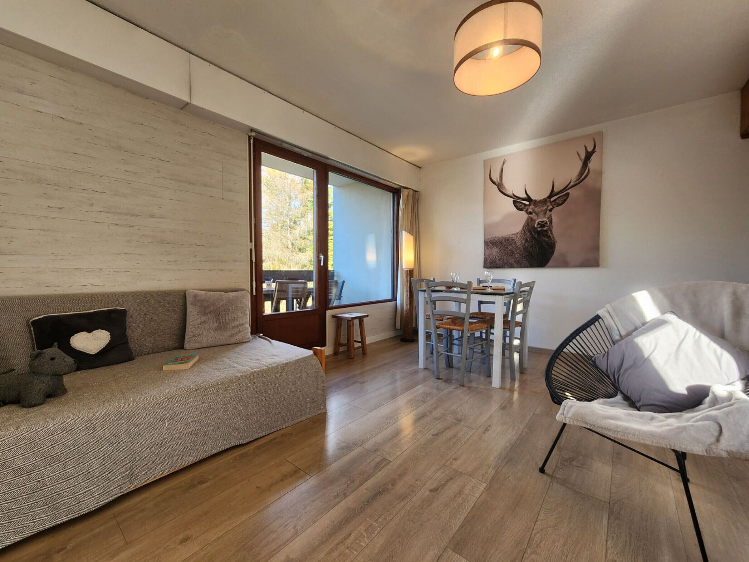 Een moderne woonkamer met een bank, een stoel, een eettafel met stoelen, een groot raam en een plafondlamp.