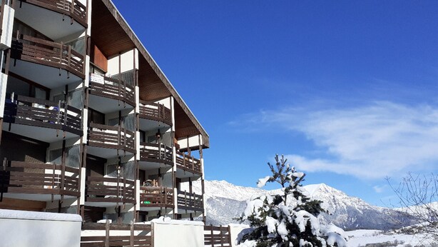 Un edificio de varios pisos con balcones, un árbol cubierto de nieve y un paisaje montañoso de fondo.