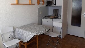 Una cocina compacta con microondas, estufa y refrigerador pequeño. Hay un estante de madera sobre la estufa y una silla en la mesa.