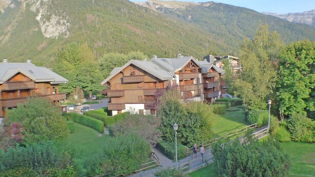 A cluster of wooden chalet-style buildings with balconies, surrounded by lush greenery and mountains in the background. - Apartment for 5 with private balcony and parking in Samoëns (Samoëns)