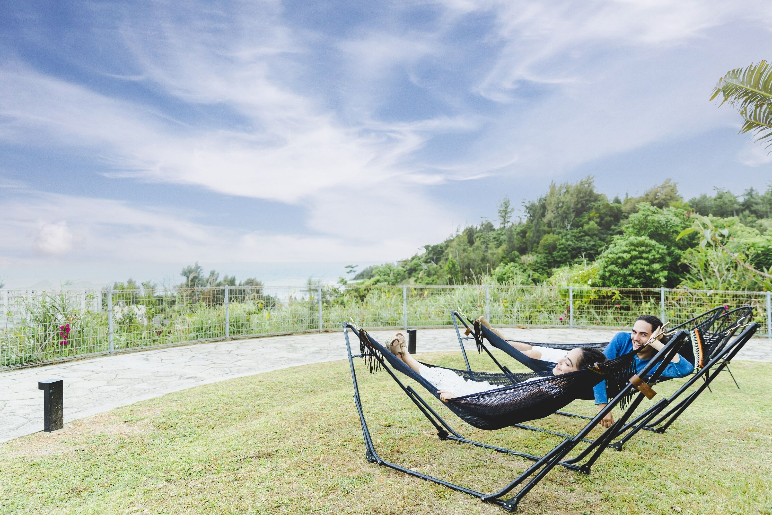 Two people relaxing in a hammock on a grassy area with a view of a lush green hill and a body of water in the background.