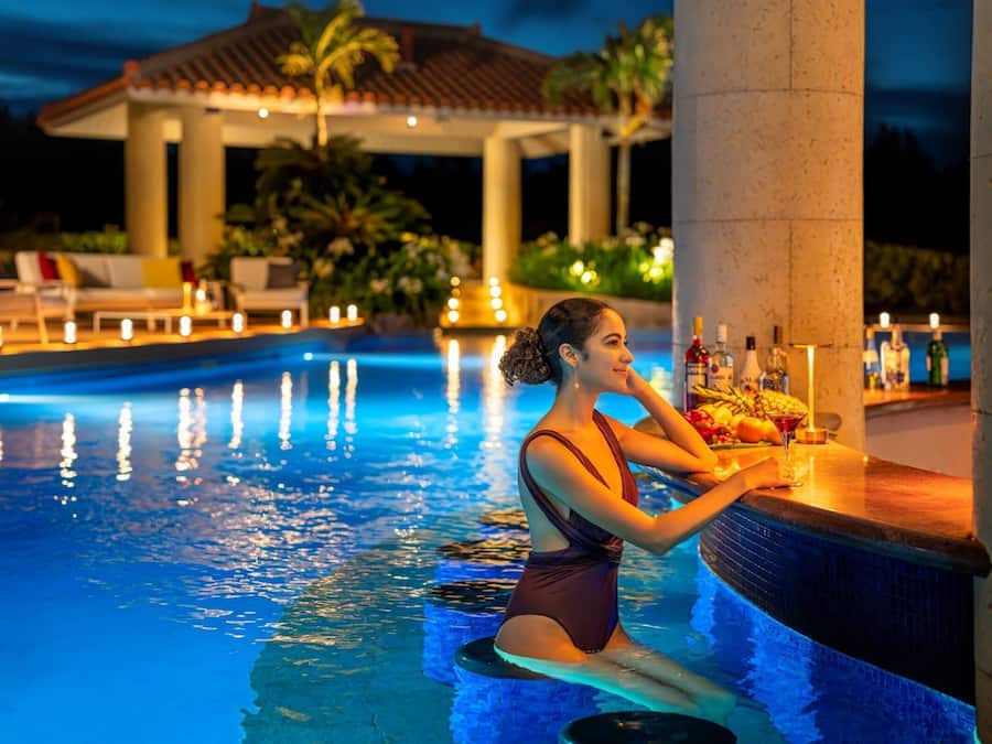 Una persona sentada junto a la barra de la piscina, con una bebida, rodeada de sillones de descanso y un pabellón al fondo.