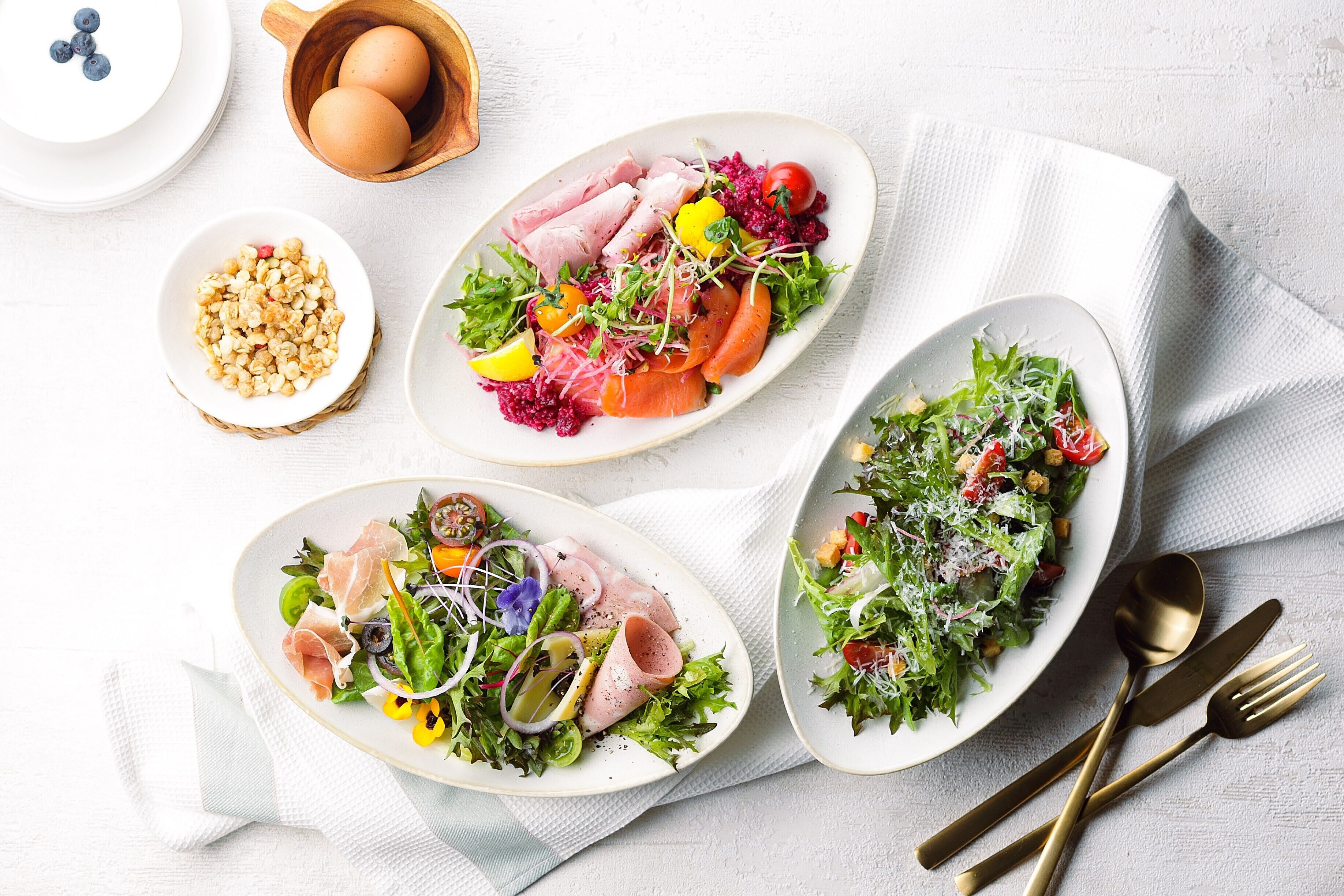 Three oval plates of food on a white tablecloth with a napkin and cutlery.