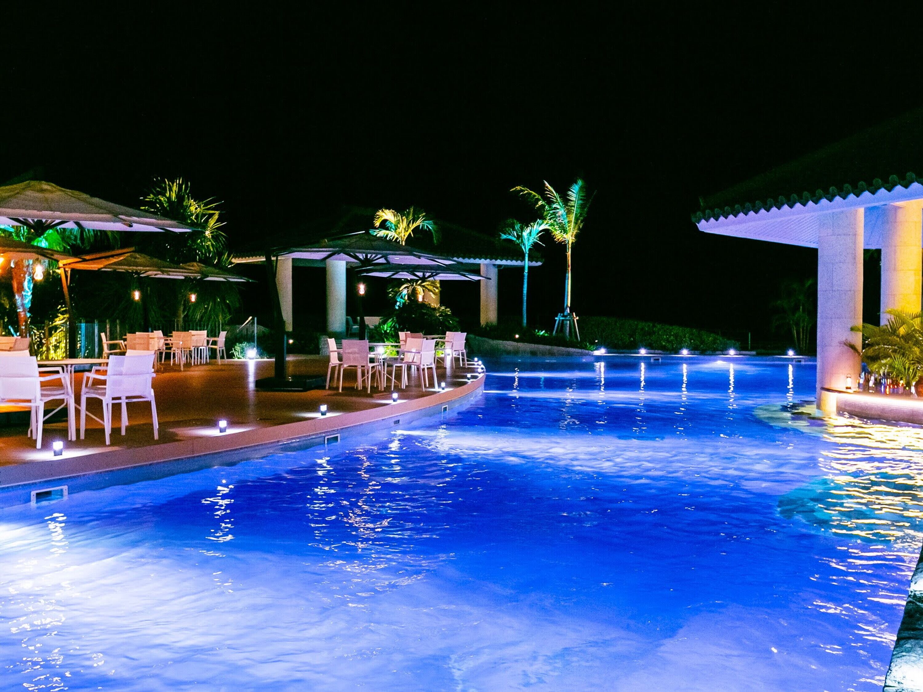 A nighttime swimming pool area with lounge chairs and umbrellas, surrounded by palm trees and a covered seating area.