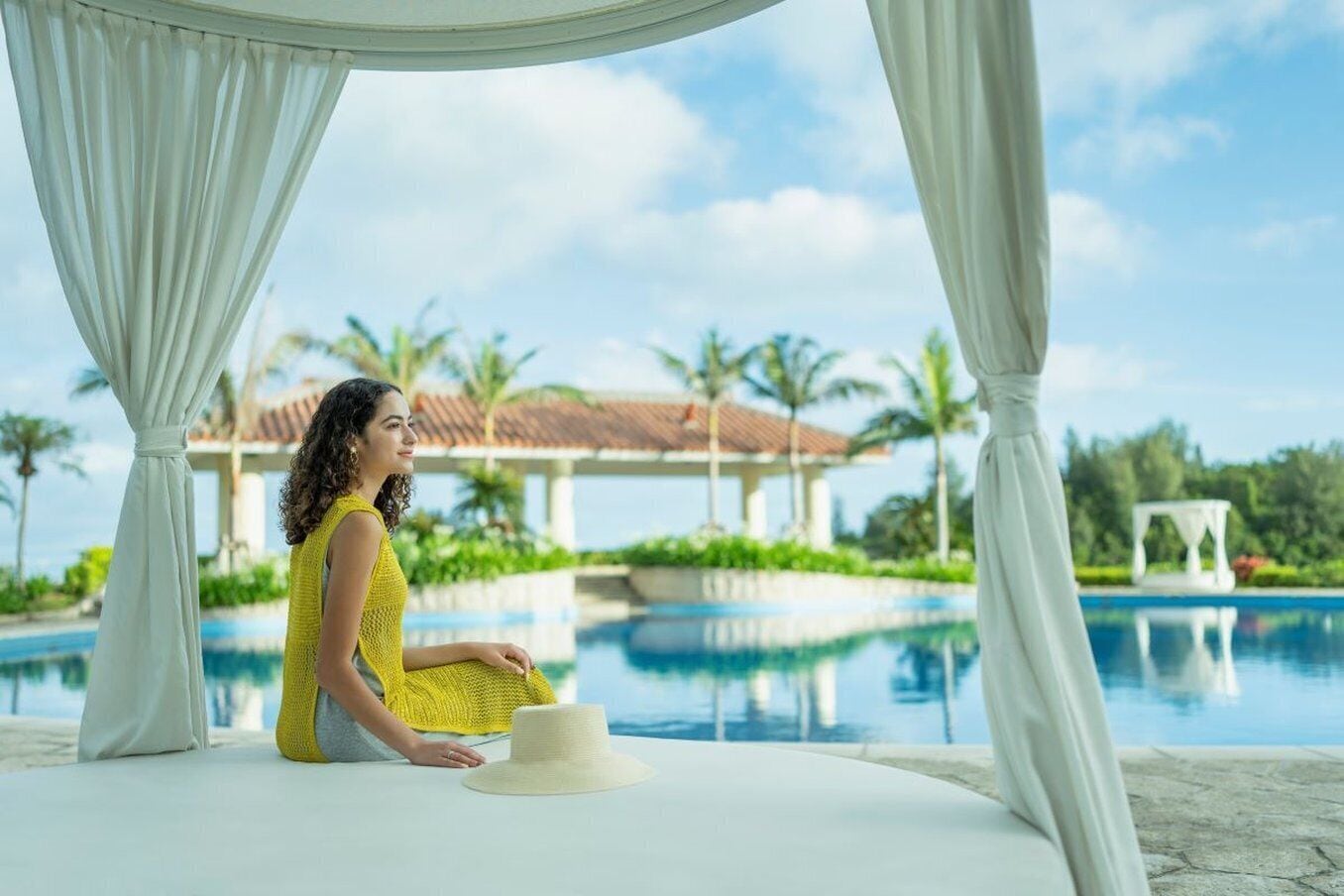 A person sitting by a pool with a white canopy, palm trees, and a building in the background.