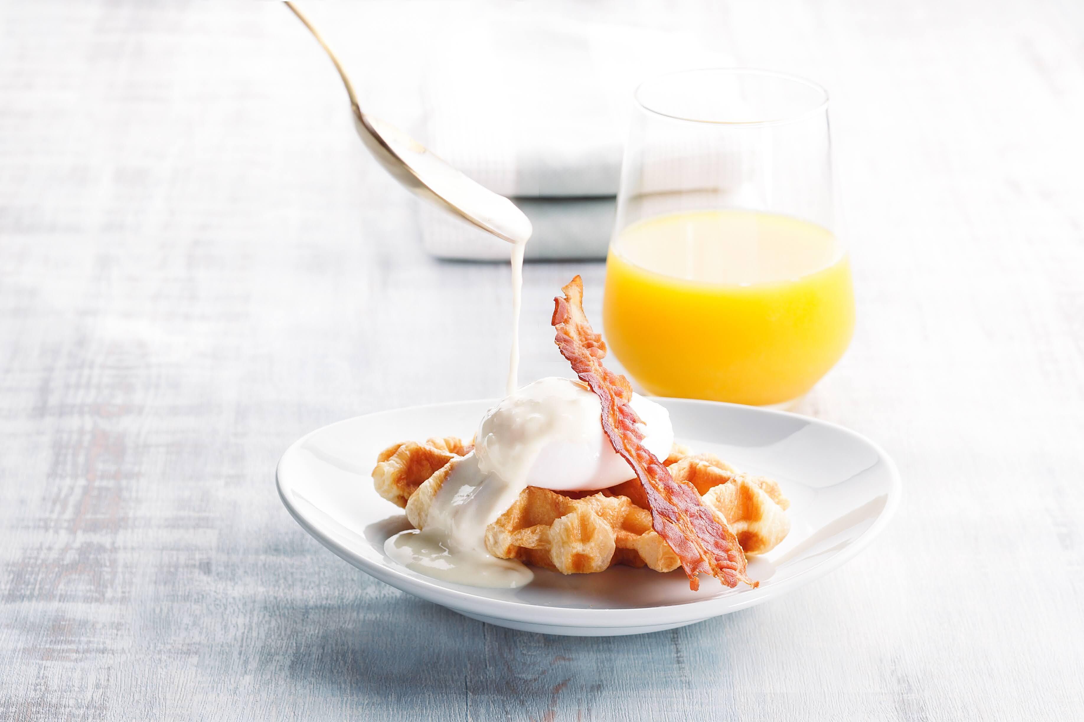 En tallrik med våfflor, ett pocherat ägg och bacon, med ett glas apelsinjuice i bakgrunden.