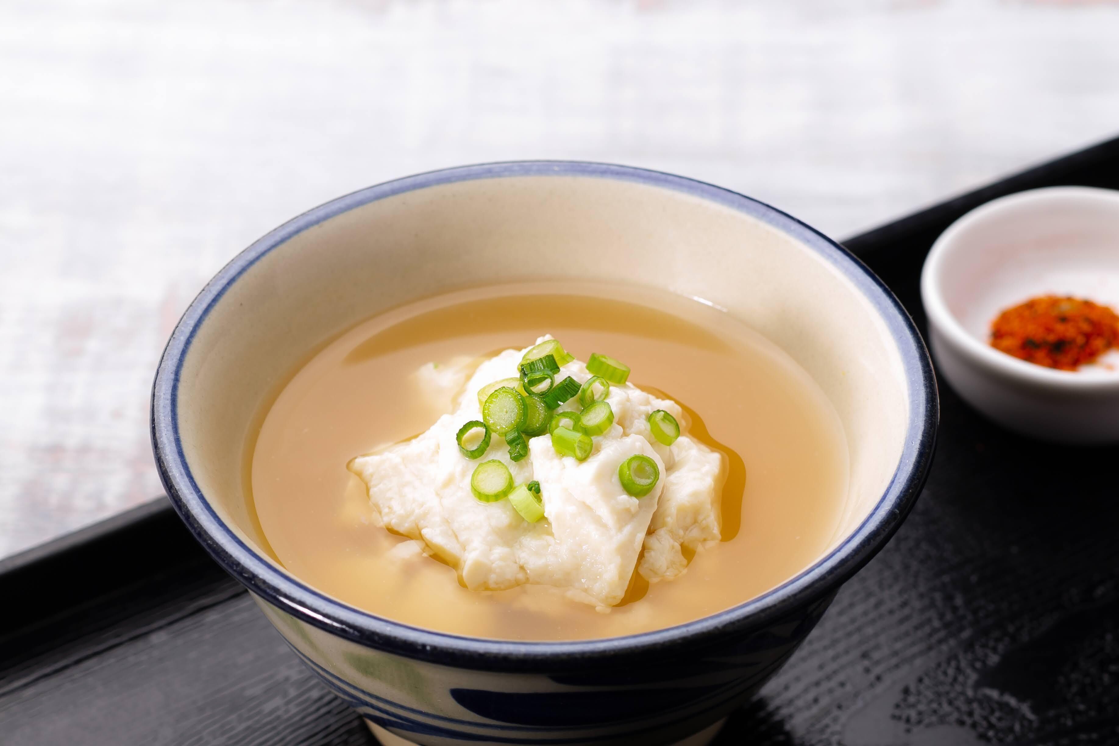 A bowl of tofu soup garnished with green onions, accompanied by a small bowl of red chili flakes.