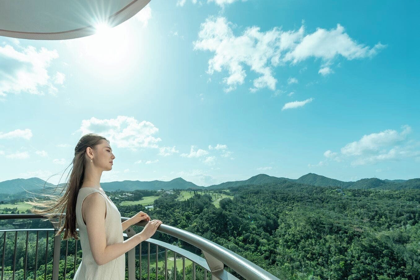 A person standing on a balcony with a view of a forested landscape and mountains.