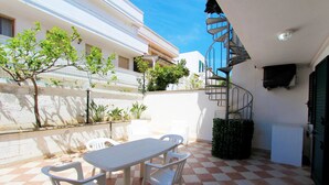 A patio with a checkered floor, a table, and chairs. There is a spiral staircase and a small garden area with plants.
