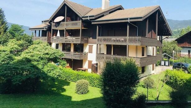Un chalet de dos pisos con balcón, rodeado de exuberante vegetación y montañas al fondo.