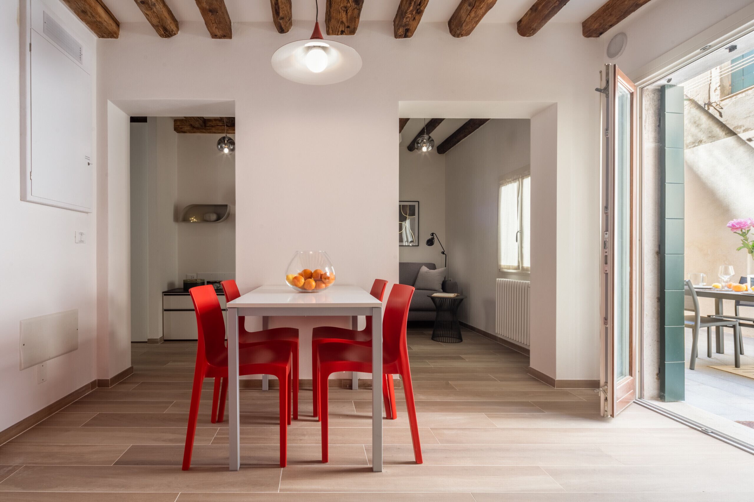 A dining area with a white table and red chairs, a bowl of fruit on the table, and a wooden ceiling.