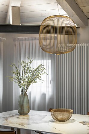 Ett matbord med en vas och en flätad skål, en hängande lampa och ett fönster med persienner.