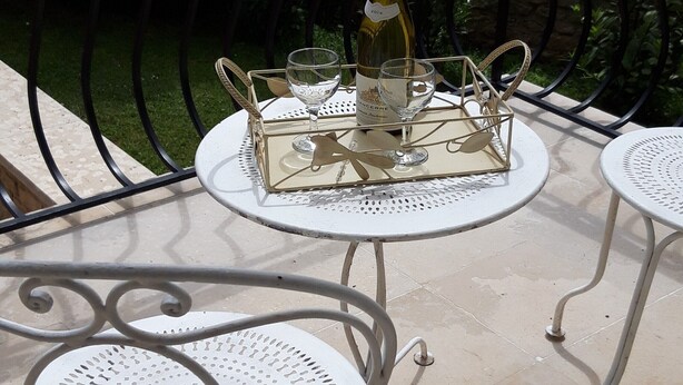 Eine Terrasse mit weißen Metallstühlen und einem Tisch, eine Weinflasche und ein Glas auf dem Tisch.
