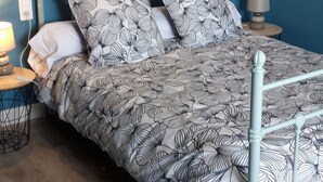Ein Schlafzimmer mit blauer Wand, einem Metallbettgestell und einer gemusterten Bettdecke. Ein Nachttisch mit Lampe und Steckdose ist vorhanden.