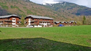 A row of wooden mountain lodges with balconies, set against a backdrop of a green field and a mountain range. - 6-Person Apartment with Terrace, Parking, and Wi-Fi (Samoëns)