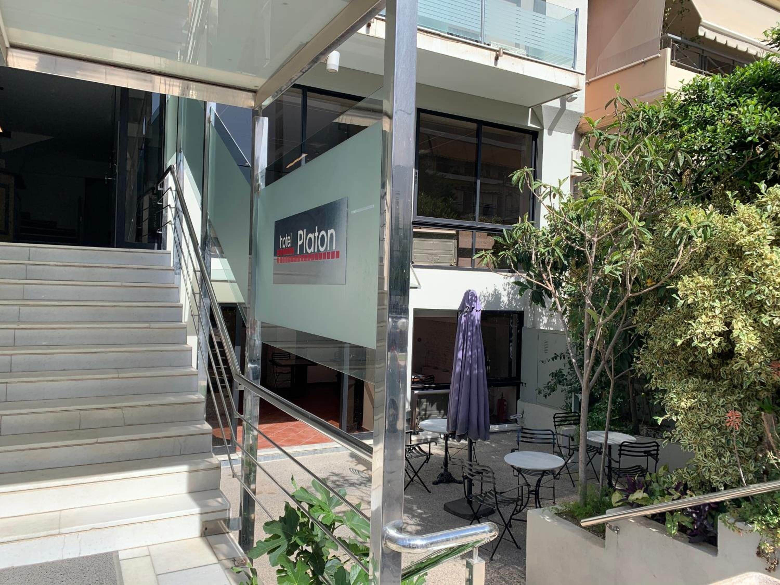 A hotel entrance with a staircase leading to the lobby, outdoor seating area with tables and chairs, and a sign for the hotel named "Platon".