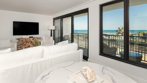 A modern hotel room with a white sofa, a flat-screen TV, a marble table, and a balcony with a view of the ocean.