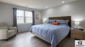Ein Schlafzimmer mit einem großen Bett, einem Sessel, einem Nachttisch und einem Fenster mit Vorhängen.