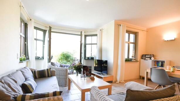 A cozy living room with a sofa, a coffee table, and a TV. There are wicker chairs and a dining table with chairs.