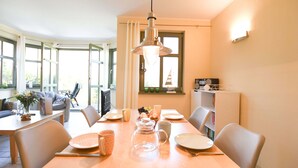 A dining area with a wooden table set for four, surrounded by chairs. There is a hanging light fixture above the table, and a window with curtains to the left.