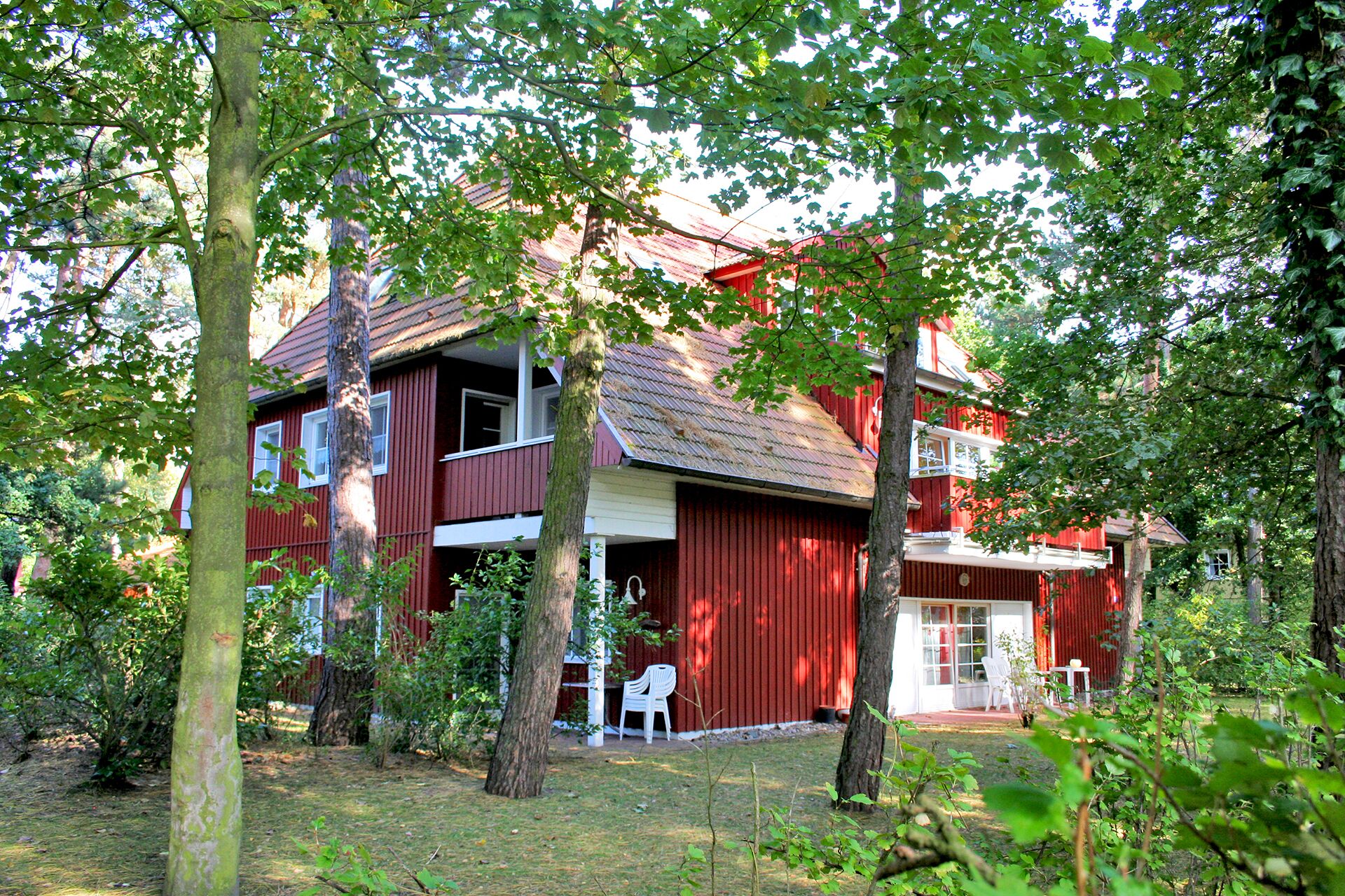 A red wooden house with a porch and white chairs, surrounded by trees.