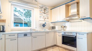 Uma cozinha com armários claros, micro-ondas, forno, pia e janela com vista para uma casa azul.