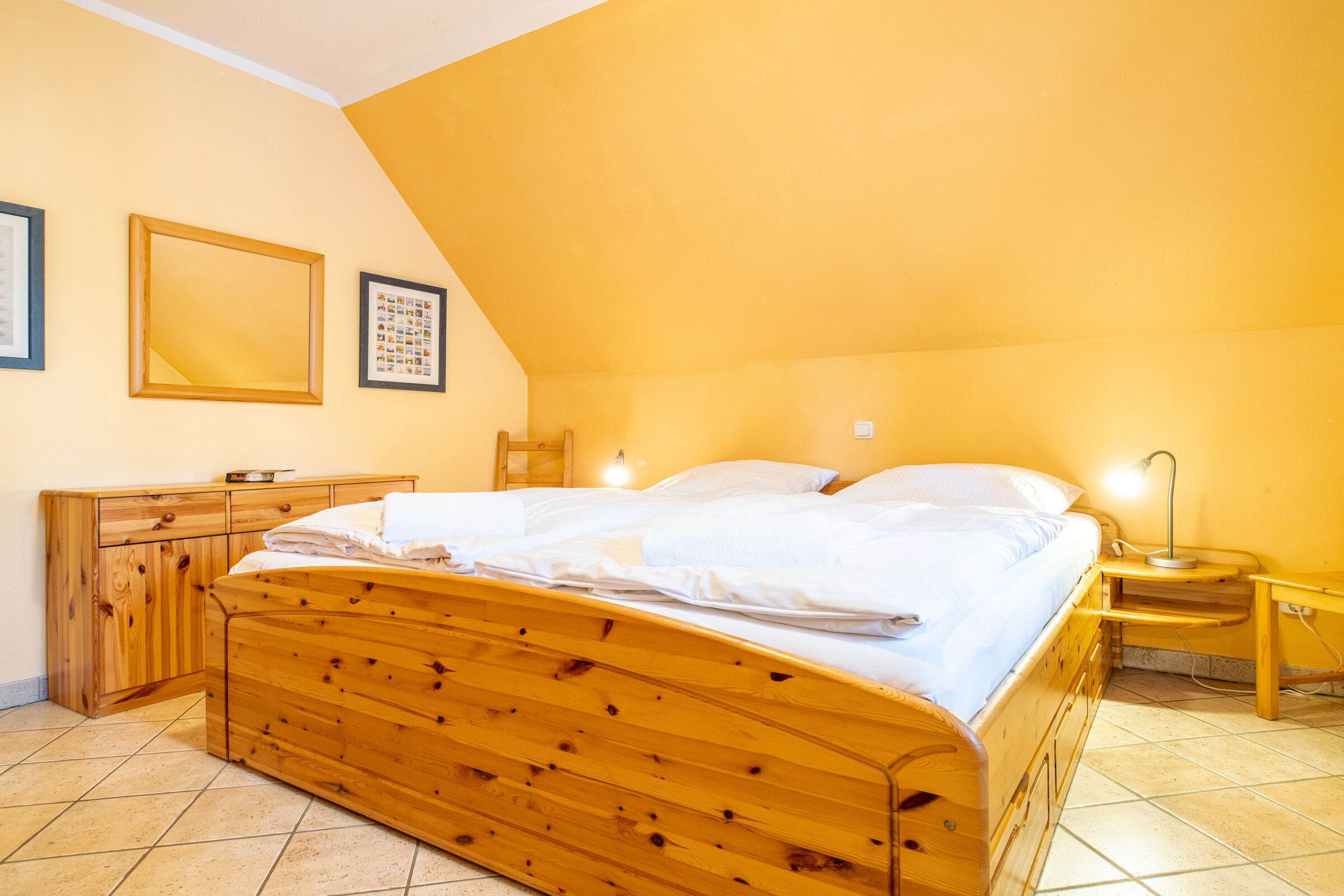 Een slaapkamer met een houten bed, een nachtkastje, een dressoir en twee ingelijste foto's aan de muur.