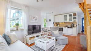 Ein modernes Wohnzimmer mit Fernseher, Sofa und Couchtisch, eine Küche mit weißen Schränken und ein Essbereich.