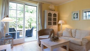 A cozy living room with a sofa, armchair, and a table with a decorative centerpiece. A cabinet with glass doors is visible, and a window offers a view of greenery outside.