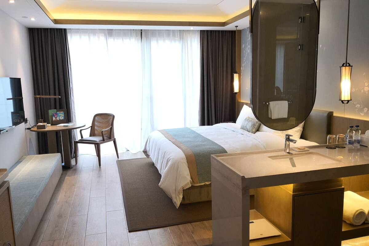 Een moderne hotelkamer met een groot bed, een bureau met stoel, een ronde spiegel en een badkamer met wastafel en handdoekhouder.