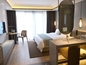 A modern hotel room with a large bed, a desk with a chair, a round mirror, and a bathroom with a sink and towel rack.