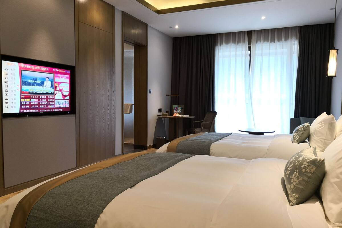 Een hotelkamer met twee bedden, een televisie aan de muur, een bureau met een stoel, en een raam met gordijnen.