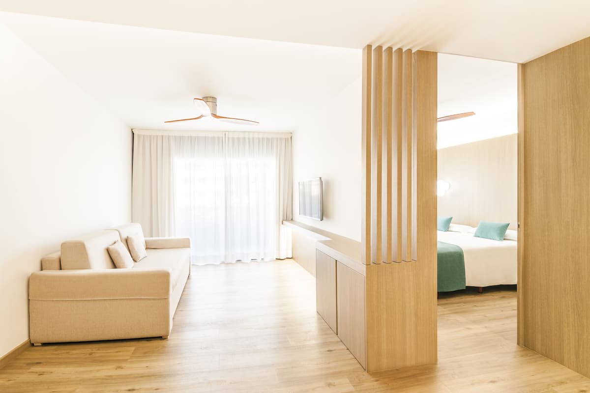 A modern, minimalist living room with a sofa, a wooden cabinet, and a bed in the background.