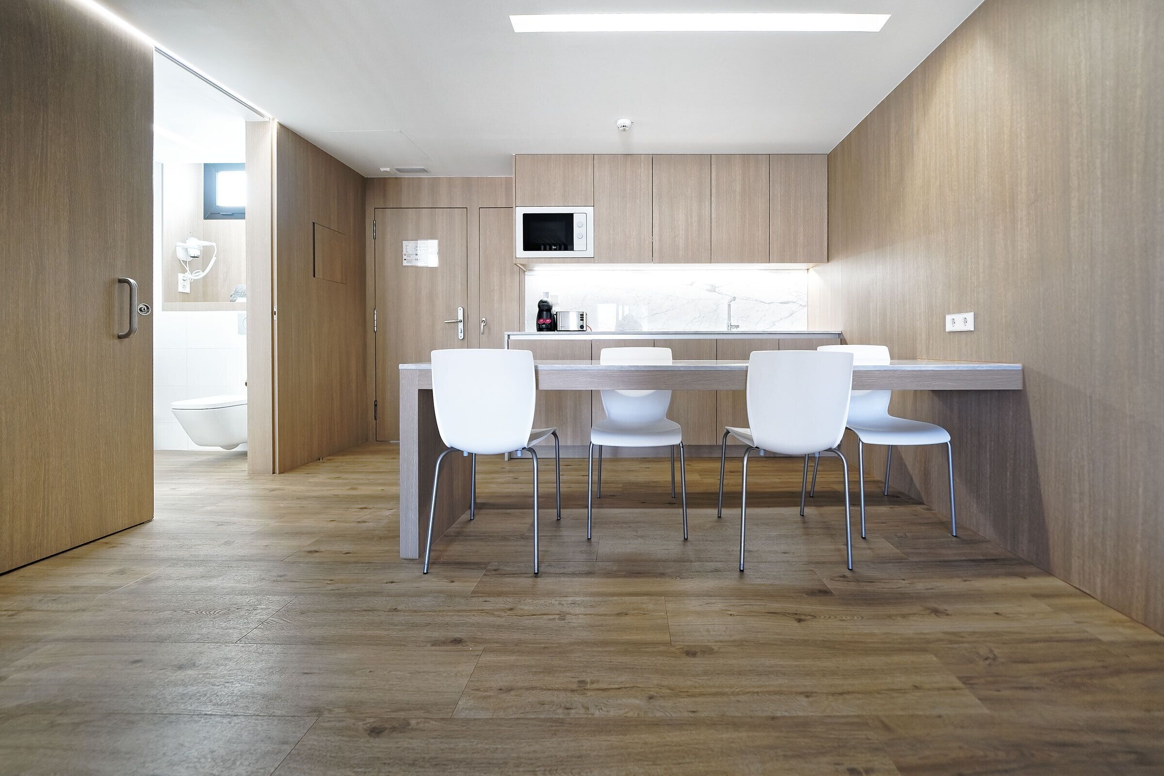 A modern kitchen with wooden cabinets, a microwave, and a dining area with white chairs.