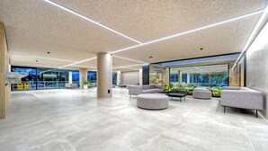 Un vestíbulo espacioso con diseño moderno, que cuenta con un amplio salón con otomanas y sofás, un techo alto con iluminación empotrada y grandes ventanales con vistas al exterior.
