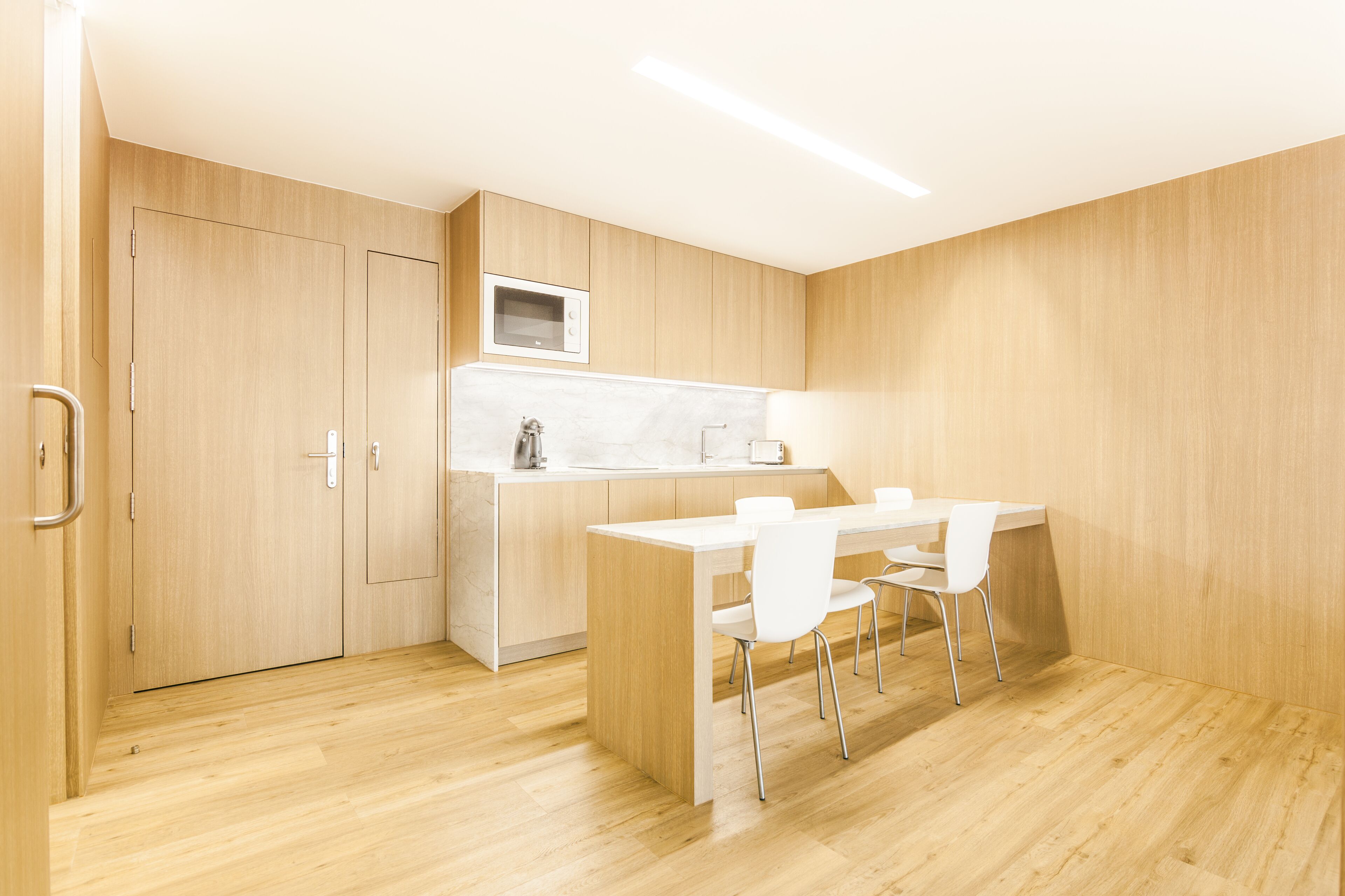 A modern kitchen with wooden cabinetry, a white countertop, and a built-in microwave. There is a dining area with a white table and chairs.