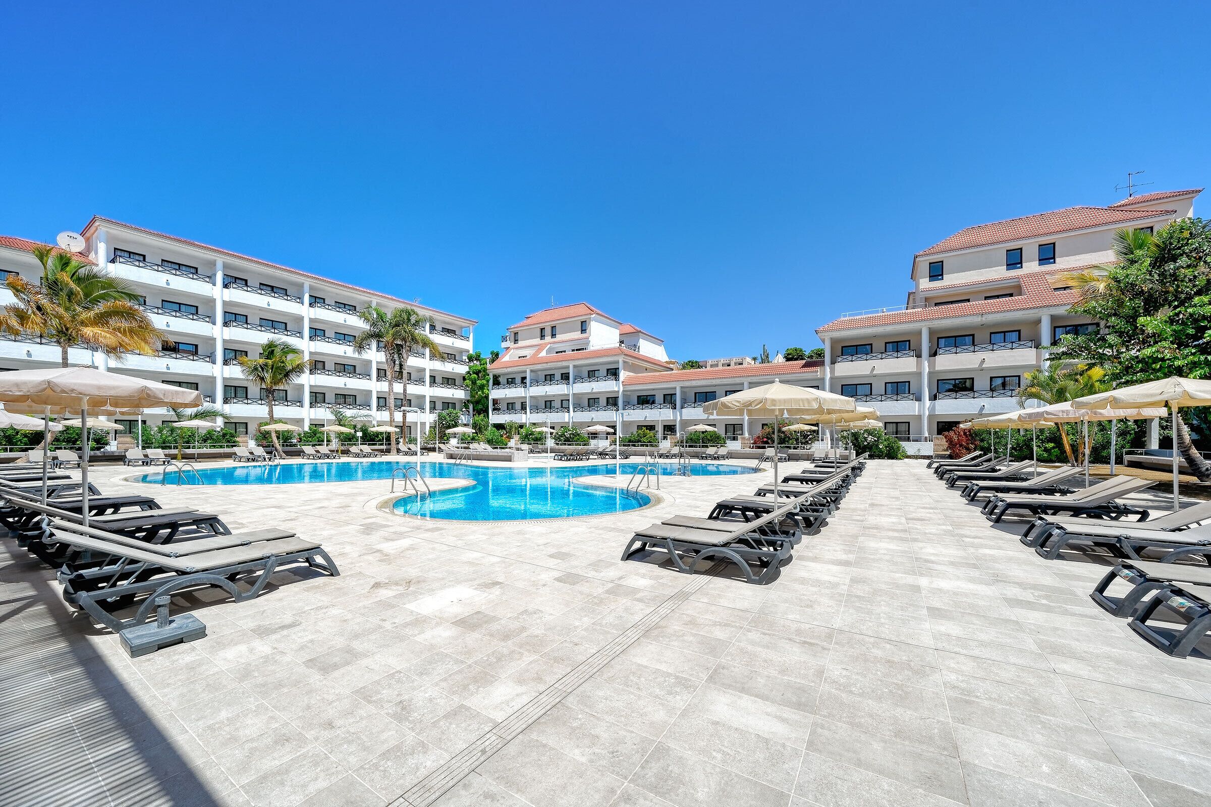 A hotel pool area with sun loungers, umbrellas, and a clear blue sky.