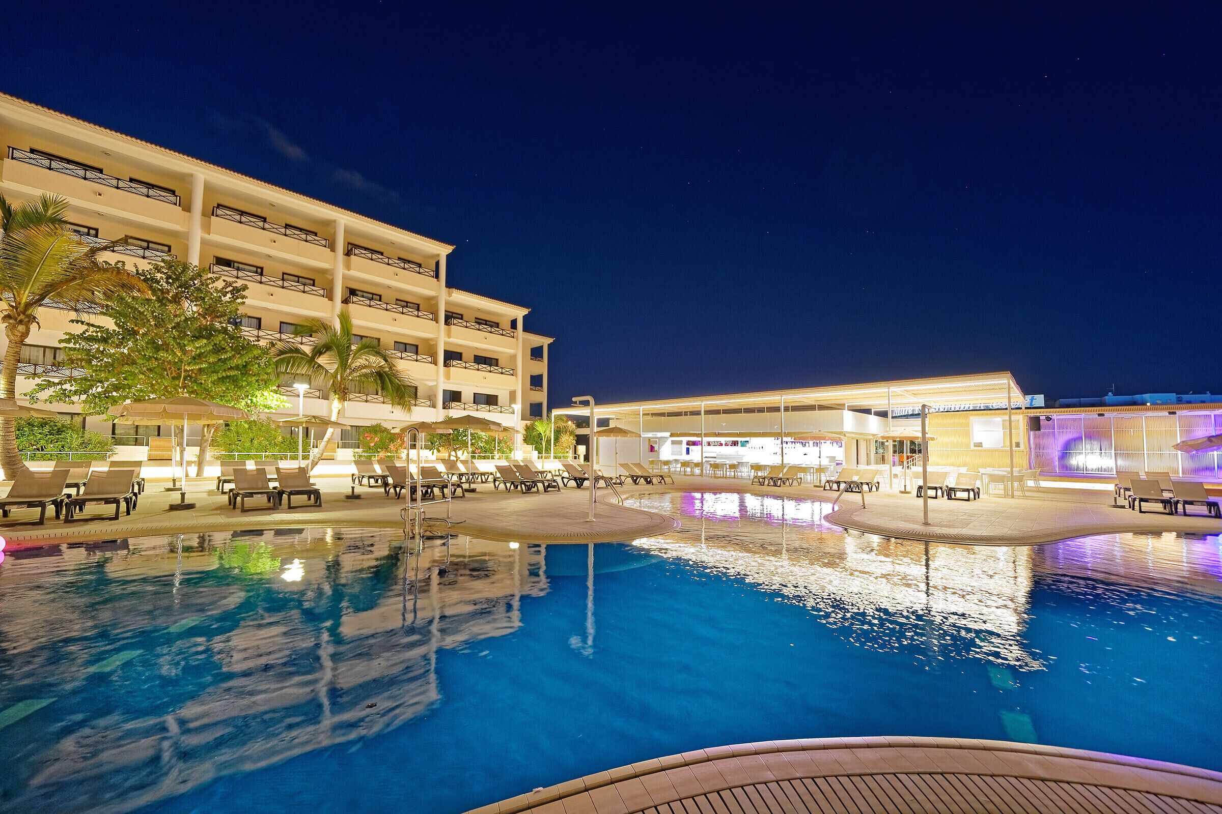 Un espace piscine d’hôtel avec des chaises longues et des parasols, entouré d’un immeuble à plusieurs étages doté de balcons.